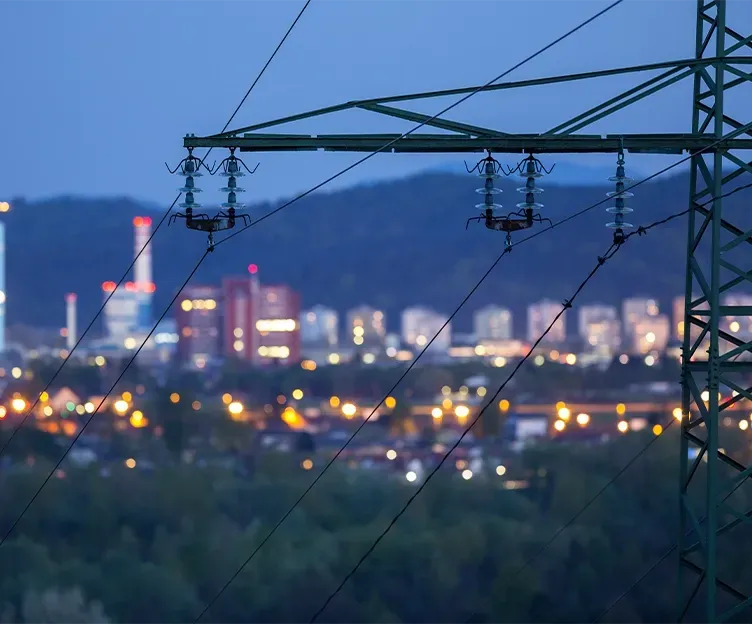 Image of transmission lines in front of a twinkling cityscape at dusk. Energy market analysis production cost analysis.